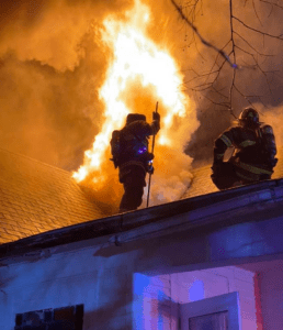 Fire fighters stand on a roof next to a blaze coming out the roof